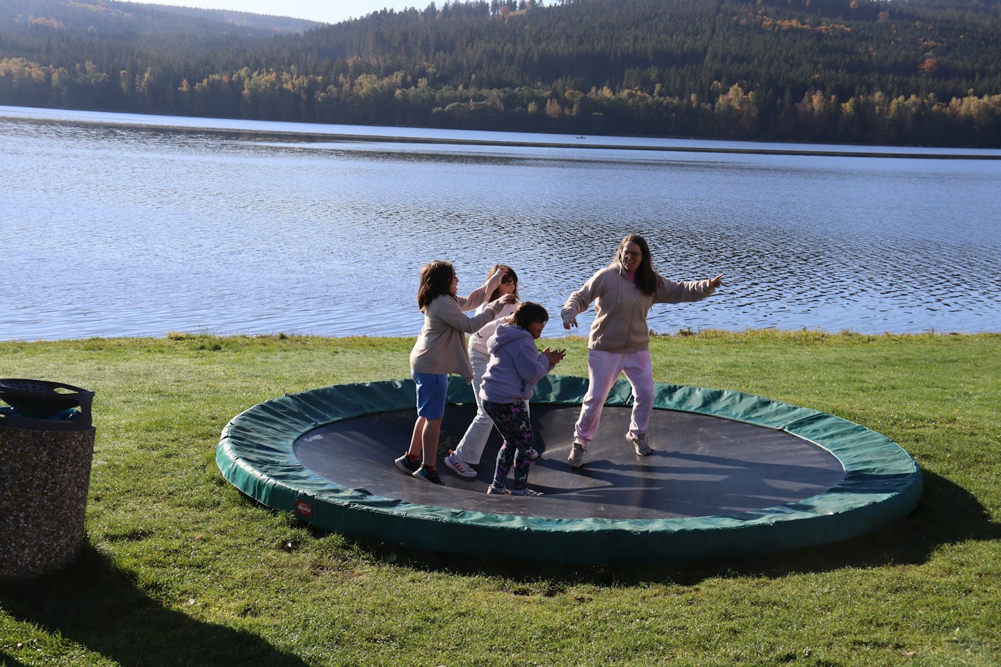 Woman and three children jump on a trampoline by a lake.